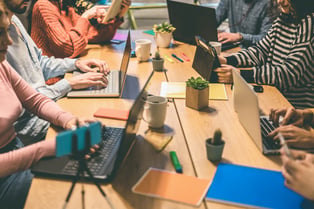 A collaborative office table is lined with gen z workers using laptops.