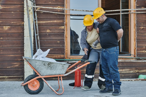 A man wearing a hard hat holds his back with his left hand while a woman wearing a hard hat assists him by holding him up with his right hand. They are at a construction site with a wheelbarrow and other equipment in the photo.